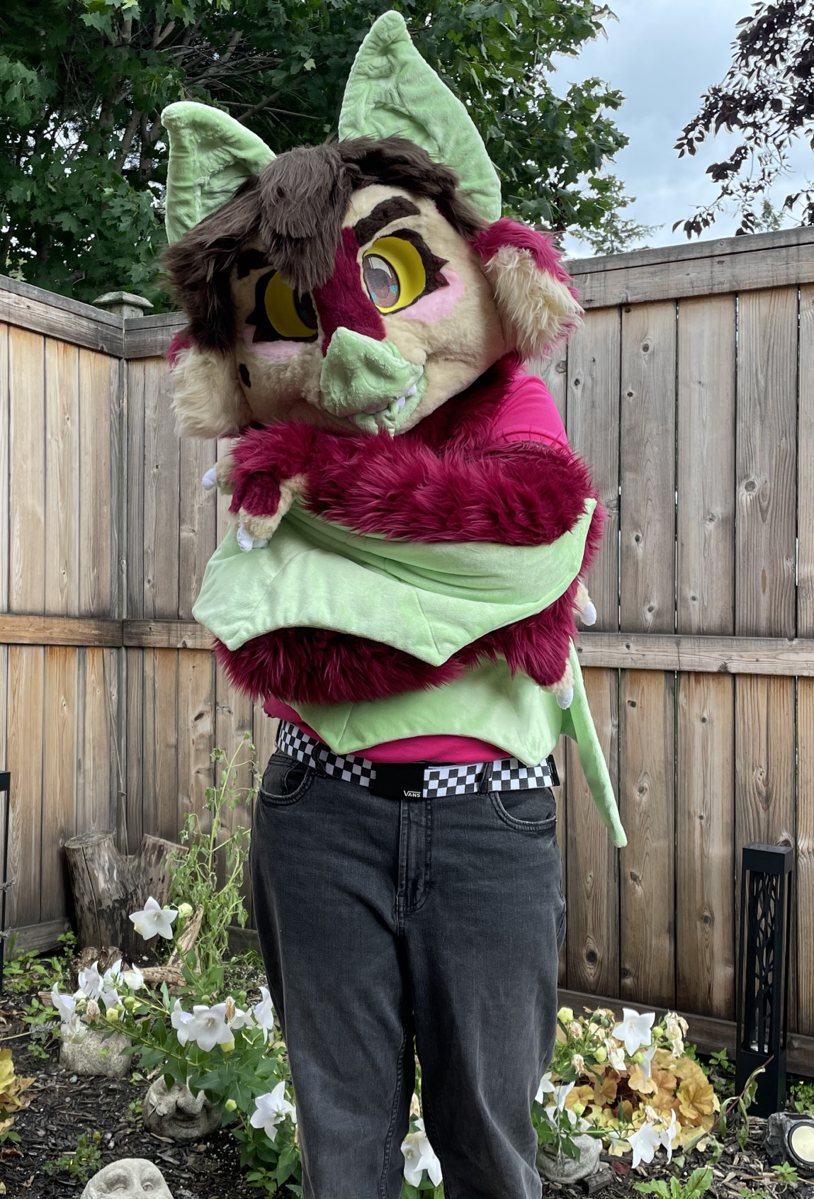 The bat fursuiter with his wings wrapped around himself. He is wearing a pink shirt, black jeans, rainbow-coloured shoes, and a black and white checkered belt