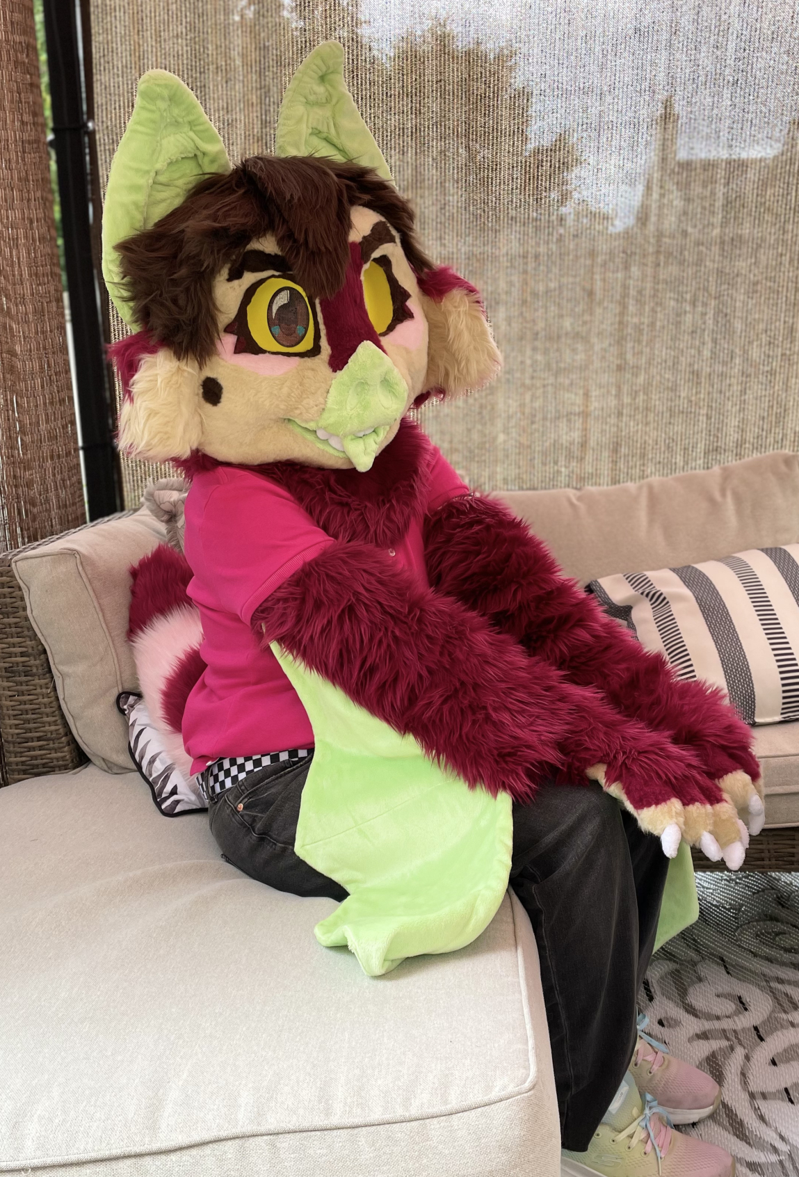 The bat fursuiter sitting on an outdoor couch, with his arms resting on his knees