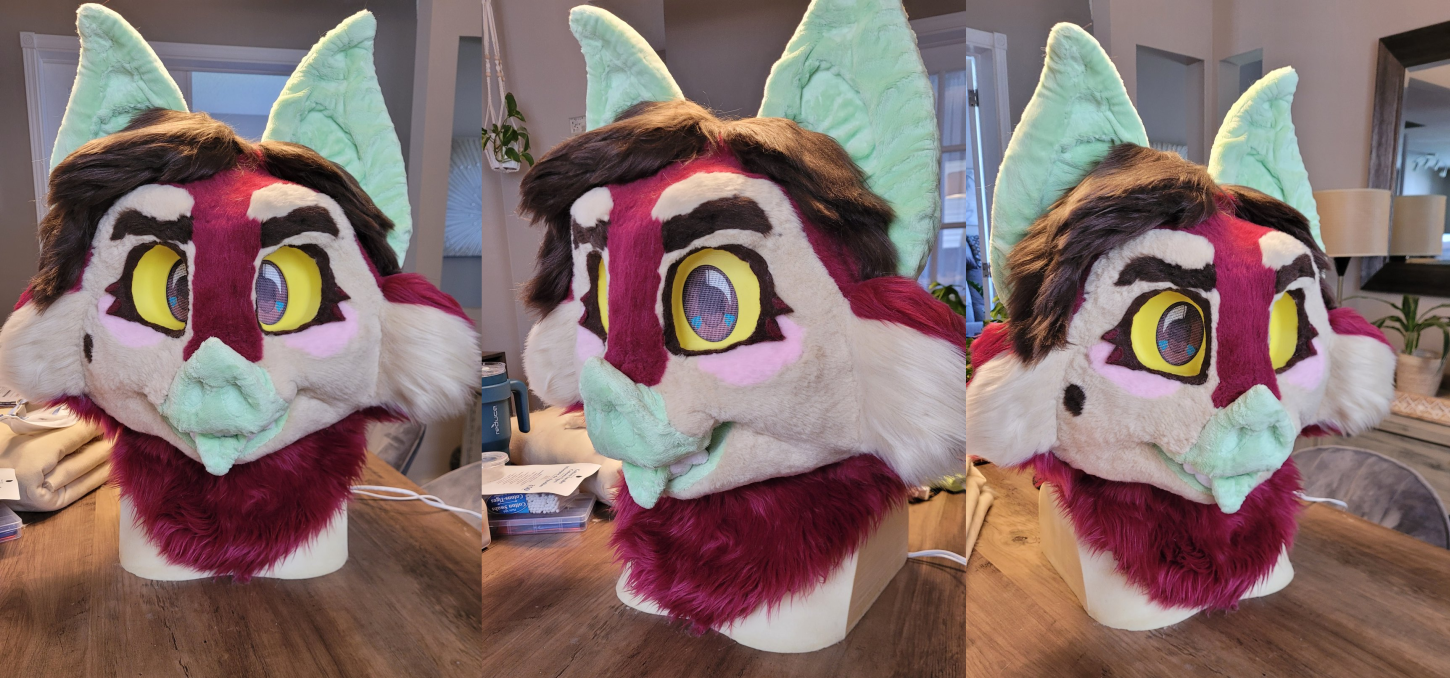 Fursuit head of a vampire bat with red, pink, and tan fur, brown hair, and green skin